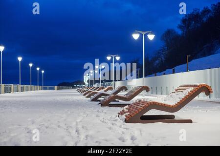 Panchine in legno alla luce delle lanterne nel parco. Parco cittadino illuminato di notte. Passerella coperta di neve. Blu scuro notte cielo nuvoloso, winte neve atmosferica Foto Stock