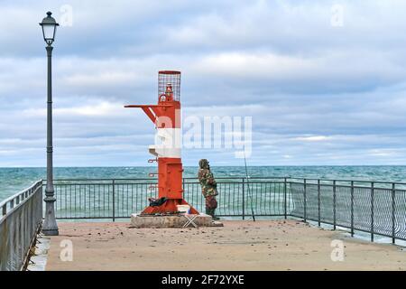 Pescatore maschile in camouflage vestito in piedi e la pesca sul molo vicino al faro rosso e bianco. Concetto di pesca in mare. Foto Stock