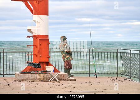 Pescatore maschile in camouflage vestito in piedi e la pesca sul molo vicino al faro rosso e bianco. Concetto di pesca in mare. Foto Stock