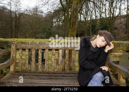 Una giovane donna attraente in un parco pubblico sedette sopra una vecchia panchina in una giornata fredda in inverno il tempo è sconvolto e spaventato Foto Stock