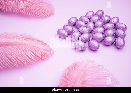 Sfondo rosa pasquale con uova di pasqua al cioccolato avvolte in lamina di colore viola con piume colorate su uno sfondo rosa chiaro. Concetto di Pasqua. AE Foto Stock