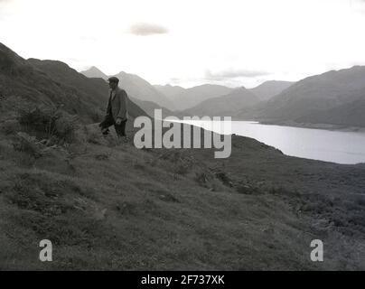 1956, storico, un uomo che indossa un cappuccio di stoffa e giacca sportiva camminando nelle alture delle Highlands scozzesi, nella Scozia nordoccidentale, con le catene montuose che circondano un grande lago o un'insenatura oceanica sottostante. L'area ospita molti laghi d'acqua grandi e profondi, noti come laghi, il famoso è "Loch Ness" e la regione montuosa ha la vetta più alta del Regno Unito, "ben Nevis". Bisogna fare molta attenzione quando si cammina o si cammina nelle alture perché il tempo può cambiare - peggio - molto rapidamente. Questa regione remota è la più umettista del Regno Unito. Foto Stock