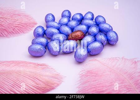 Sfondo rosa pasquale con uova di pasqua al cioccolato avvolte in lamina di colore viola con piume colorate su uno sfondo rosa chiaro. Concetto di Pasqua. AE Foto Stock