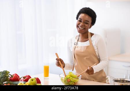 Eccitata Africana Female che fa insalata vegetale in piedi in cucina moderna Foto Stock