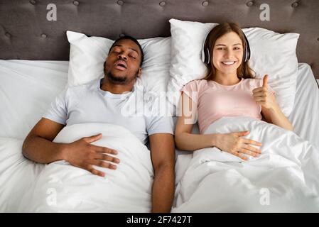 Felice giovane donna che indossa le cuffie e mostra il pollice in su gesto mentre suo marito russare sul letto, vista dall'alto Foto Stock