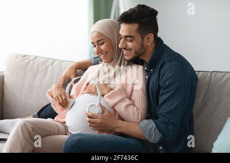 Musica di gravidanza. Felice coppia musulmana incinta che mette le cuffie senza fili sul ventrido della donna Foto Stock