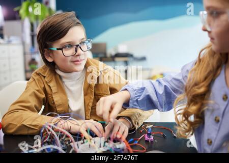 Ritratto di ragazzo e ragazza costruendo robot e sperimentando con circuiti elettrici durante la classe di ingegneria a scuola, copia spazio Foto Stock