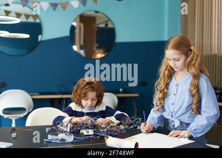 Ritratto di schemi di disegno di ragazzo e ragazza durante la costruzione di robot durante la lezione di ingegneria a scuola, copia spazio Foto Stock