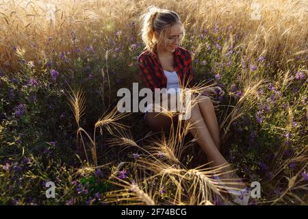 Una giovane donna al tramonto si siede in un campo con fiori e grano. Foto Stock