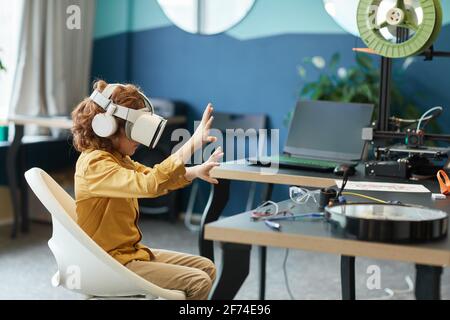 Ritratto in vista laterale di un bambino che indossa un visore VR e che si sta sportando fuori mentre prova la tecnologia aumentata nel laboratorio scolastico, spazio di copia Foto Stock