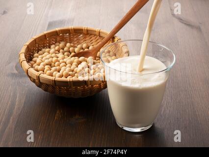 Versare il latte di soia in un bicchiere posto su uno sfondo di legno. Fagioli di soia in un colander di bambù sullo sfondo Foto Stock