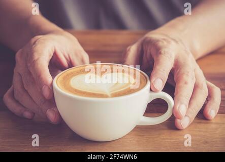 Mani di un uomo o barista che tiene una tazza di caldo latte di caffè con schiuma a forma di cuore art Foto Stock