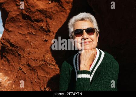 Felice anziana turista femminile in abbigliamento casual appoggiato a mano seduto su una formazione rocciosa con terreno arido sotto il blu nuvoloso cielo Foto Stock