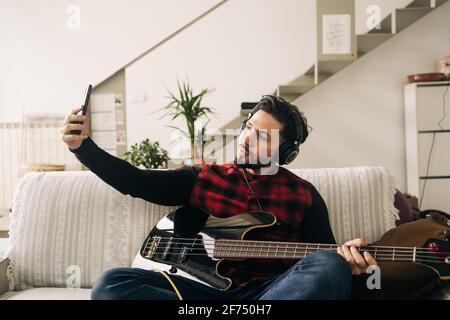 Unshaven artista maschile in cuffia con chitarra elettrica prendendo auto ritratto sul cellulare mentre si siede sul divano in casa Foto Stock