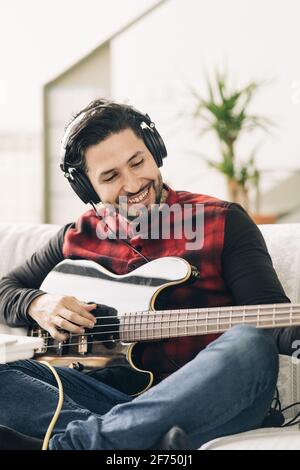 Contenuto artista maschile adulto in cuffia che suona la chitarra basso su divano nella stanza della casa nella luce del giorno Foto Stock