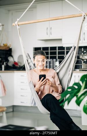 Cellulare di navigazione femminile di mezza età positivo mentre si siede in comodo amaca vicino banco cucina con utensili da cucina a casa con verde pianta Foto Stock