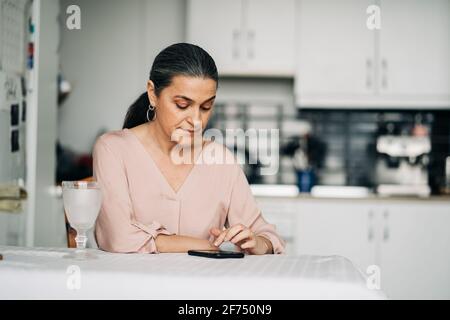 Cellulare da surf concentrato di mezza età mentre si siede a tavola in cucina moderna con utensili da cucina a casa Foto Stock