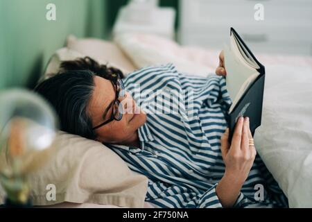Donna ispanica matura e rilassata in abiti casual e occhiali sdraiati a letto e leggere interessante libro prima di dormire Foto Stock