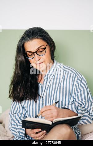 Donna allegra di mezza età in camicia a righe sorridente e scrittura notebook a letto con smartphone Foto Stock