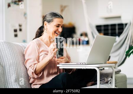 Vista laterale di una femmina sorridente di mezza età seduta sul divano e mostrare l'immagine del bambino mentre si ha la chat video su netbook su sfondo sfocato Foto Stock
