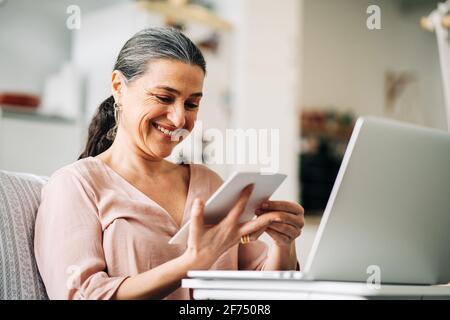 Allegra femmina di mezza età seduta sul divano vicino a un netbook moderno mentre si guarda la foto in appartamento moderno con cucina su sfondo sfocato Foto Stock