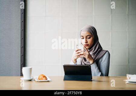 Vista laterale della donna musulmana freelance che indossa la tradizionale hijab in piedi in bar e smartphone durante la navigazione mentre si lavora a progetti in remoto Foto Stock