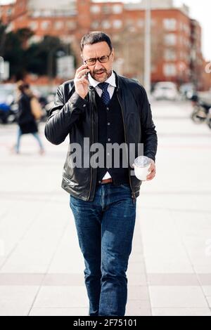 Maturo uomo ispanico executive in abbigliamento casual elegante con caldo bere per andare a parlare sul cellulare mentre si cammina in città pavimentazione Foto Stock