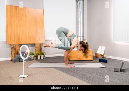Tutta la lunghezza di misura giovane donna che esegue Bakasana mentre pratica yoga sull'uomo in appartamento moderno vicino smartphone su treppiede Foto Stock