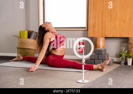 Vista laterale di flessibile barefooted Woman in elegante rosso top e leggings esecuzione di Front splits durante la registrazione di vlog su smartphone collegato al treppiede i. Foto Stock