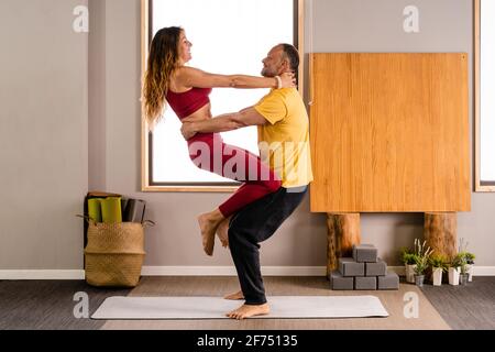 Vista laterale di coppia positiva in abbigliamento sportivo che esegue yoga asana insieme mentre si pratica lo yoga a casa alla luce del giorno Foto Stock