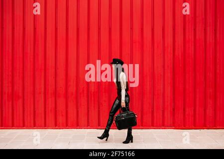 Vista laterale dell'imprenditore femminile che indossa abiti alla moda lungo la strada strada sullo sfondo di muro rosso e guardando via Foto Stock