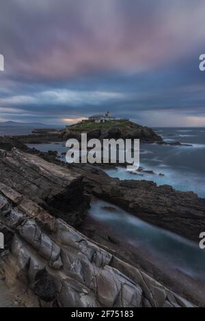 Scenario mozzafiato di isola rocciosa con faro situato in oceano Vicino costa rocciosa a Faro Tapia de Casariego nelle Asturie In Spagna sotto la nuvolosa sk Foto Stock