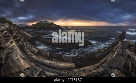 Scenario mozzafiato di isola rocciosa con faro situato in oceano Vicino costa rocciosa a Faro Tapia de Casariego nelle Asturie In Spagna sotto la nuvolosa sk Foto Stock