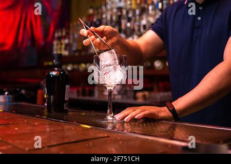 Barista irriconoscibile che mette un grande cubetto di ghiaccio nel bicchiere mentre preparate un cocktail tonico in gin al bar Foto Stock