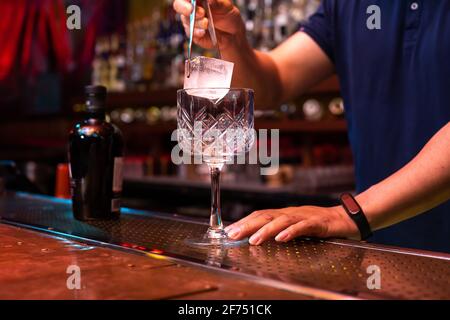 Barista irriconoscibile che mette un grande cubetto di ghiaccio nel bicchiere mentre preparate un cocktail tonico in gin al bar Foto Stock