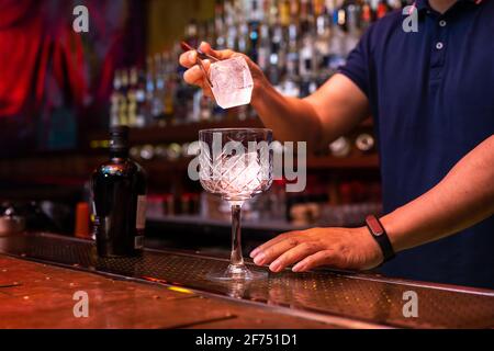 Barista irriconoscibile che mette un grande cubetto di ghiaccio nel bicchiere mentre preparate un cocktail tonico in gin al bar Foto Stock