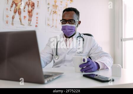 Irriconoscibile concentrato africano americano medico maschile in accappatoio medico e. maschera bere caffè e lavorare su computer portatile in clinica moderna Foto Stock