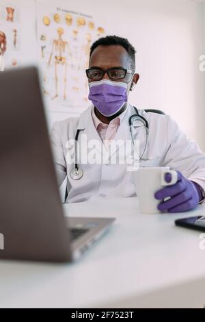 Irriconoscibile concentrato africano americano medico maschile in accappatoio medico e. maschera bere caffè e lavorare su computer portatile in clinica moderna Foto Stock