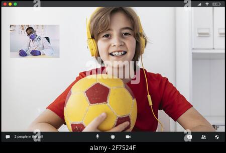 Il ragazzo è felice con le cuffie con la palla in mano che sorride mentre Avere video chat con irriconoscibile afroamericano maschio medico in accappatoio medico e mas Foto Stock