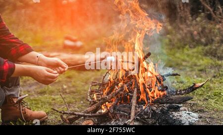 La mano della donna tiene i marshmallows dolci sul bastone sopra il falò al fuoco in foresta Foto Stock