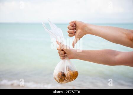 le mani tengono un sacchetto di plastica con frutta e mostrano un pollice giù sullo sfondo del mare.. nessun volto. il concetto di inquinamento della natura e di una lancetta Foto Stock