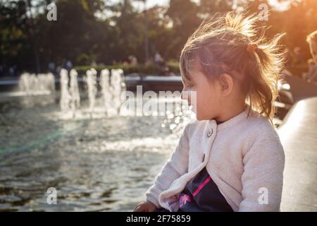 adorabile bambina guarda il laghetto con fontane nel parco in una giornata di sole. weekend a piedi in famiglia. passare il tempo con i bambini. attenzione artistica Foto Stock