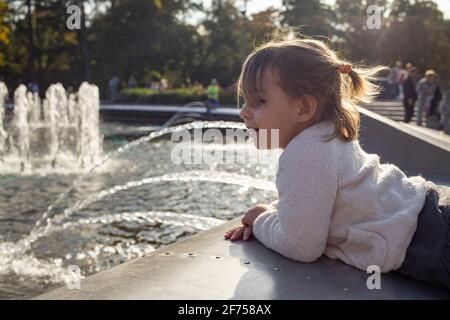 adorabile bambina guarda il laghetto con fontane nel parco in una giornata di sole. weekend a piedi in famiglia. passare il tempo con i bambini. attenzione artistica Foto Stock