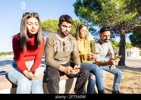 Gruppo di giovani seri seduti su una parete della città che trascorrono del tempo utilizzando uno smartphone con connessione mobile. Nuove abitudini umane normali e relatio reciproco Foto Stock