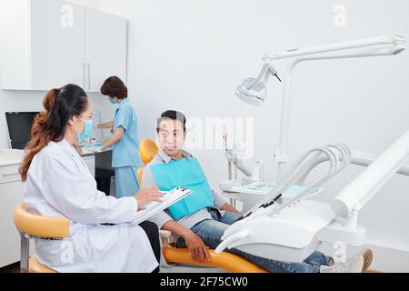 Dentista femminile che parla con il paziente e che compila il documento medico quando infermieri che preparano gli strumenti in background Foto Stock