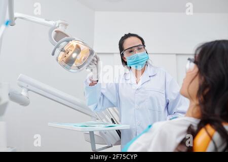 Dentista femminile esperto in occhiali protettivi e maschera medica accendere la lampada e parlare con il paziente in sedia dentale Foto Stock