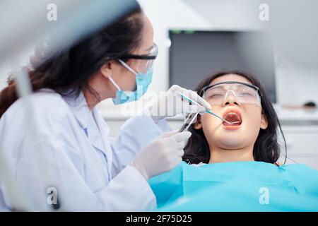 Dentista che utilizza uno specchio dentale e pinzette durante il controllo dei denti di giovane paziente femminile Foto Stock