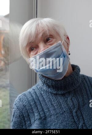 Donna anziana solitaria che indossa una maschera medica protettiva seduta vicino alla finestra a casa per la protezione dal virus. Foto Stock