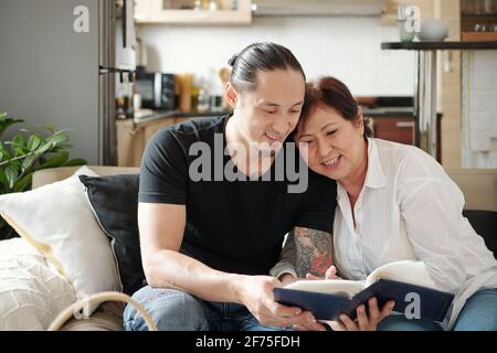 Allegra razza mista madre matura e suo figlio adulto trascorrere il tempo insieme e guardando le foto nel vecchio album fotografico Foto Stock