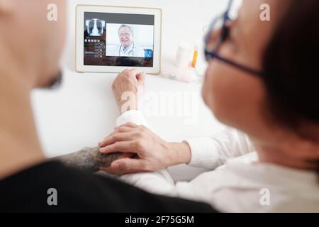 Donna anziana e suo figlio adulto seduto a tavola con tablet digitale e sessioni di telemedicina con un medico Foto Stock